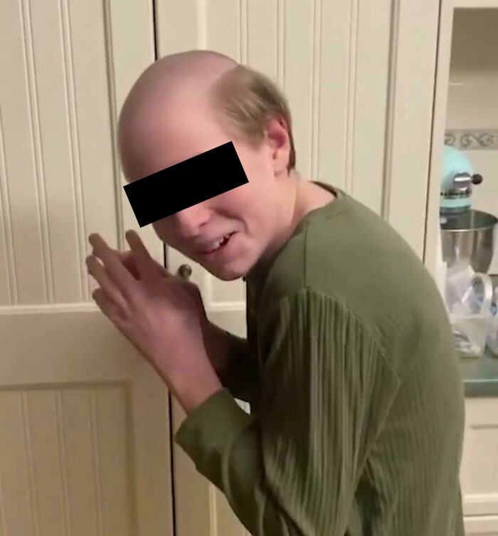 Person with a humorous haircut, wearing a green shirt, standing near a cabinet in a kitchen setting.