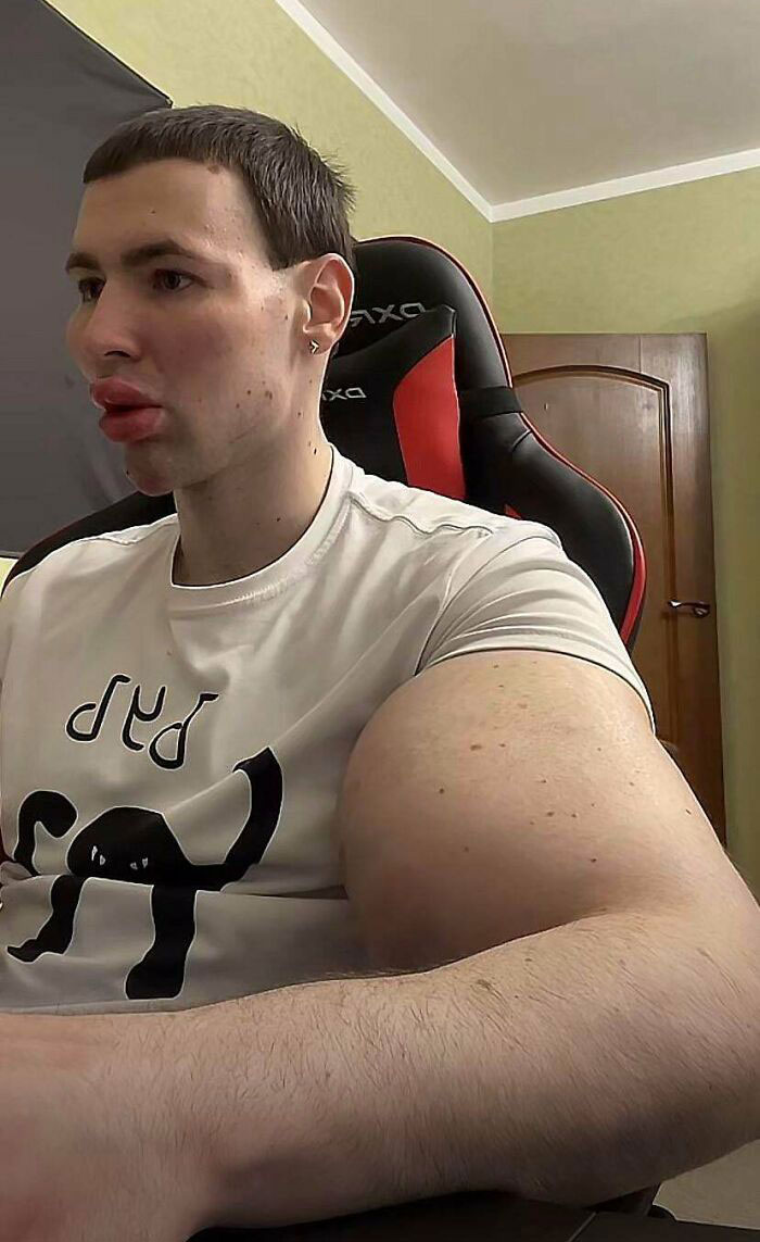 Man with a unique haircut sits in a gaming chair, wearing a white t-shirt with a black graphic design.