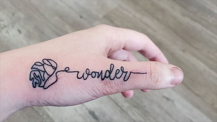 Minimalist line tattoo on hand featuring a flower and the word "wonder".