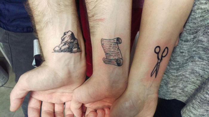 Small, simple brother and sister tattoos featuring rock, scroll, and scissors designs on forearms.