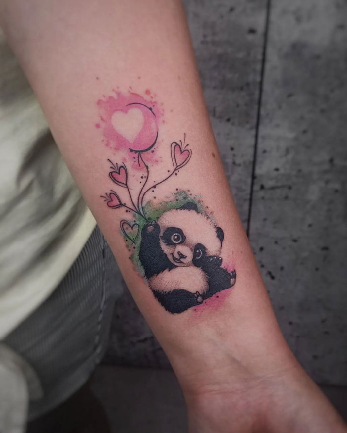Watercolor tattoo of a panda with pink heart balloons on forearm, showcasing ethereal style and vibrant colors.