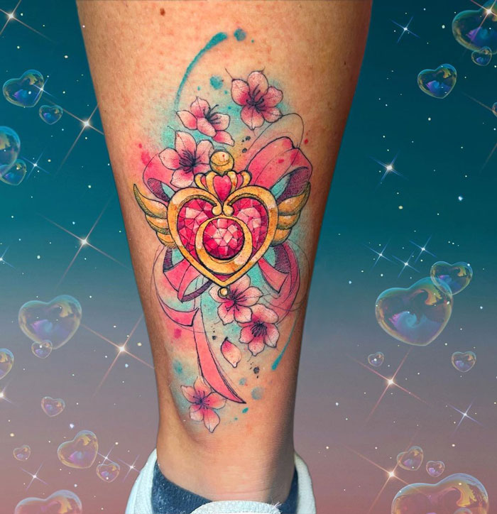 Watercolor tattoo featuring a heart with wings and pink flowers on a person's leg with a dreamy background.