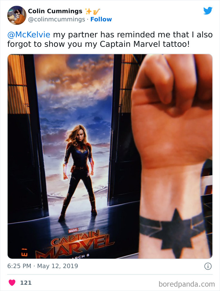 Captain Marvel tattoo on forearm with movie poster in background, showcasing superhero theme.