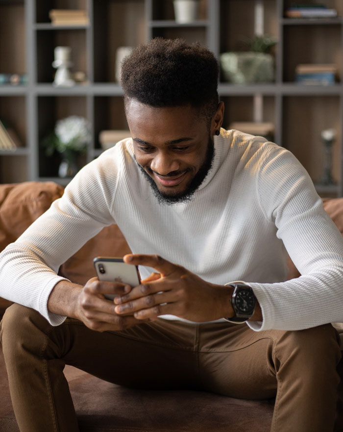 Man smiling and using a smartphone at home to avoid boredom, sitting on a sofa in a cozy living room.