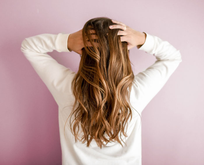 Person enjoying a relaxing moment at home, running fingers through long wavy hair, against a soft pink backdrop.