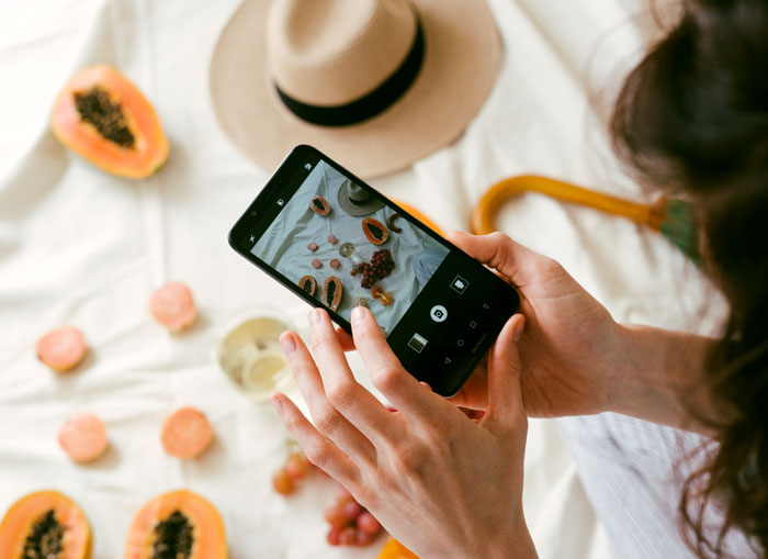 Person photographing fruits with a smartphone at home, surrounded by papaya and hat, capturing creative ways to avoid boredom.