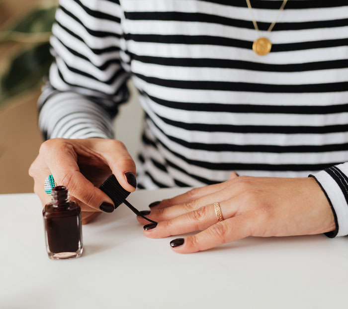 Person painting nails at home to avoid boredom, wearing a striped shirt and a gold necklace.