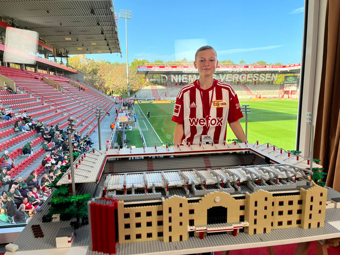 Young fan standing behind an incredible LEGO build of a detailed stadium, showcasing impressive artistic craftsmanship.