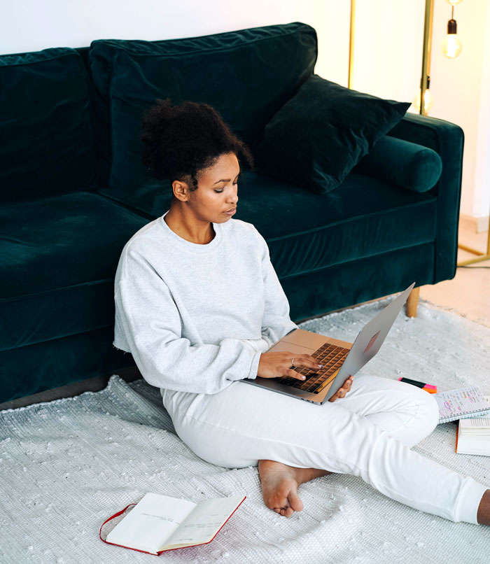Woman sitting on a rug with a laptop and a notebook, engaging in fun activities at home to avoid boredom.