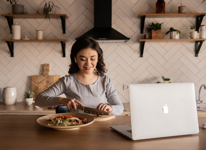 Woman enjoying fun activity at home, preparing a salad in the kitchen with a laptop nearby.