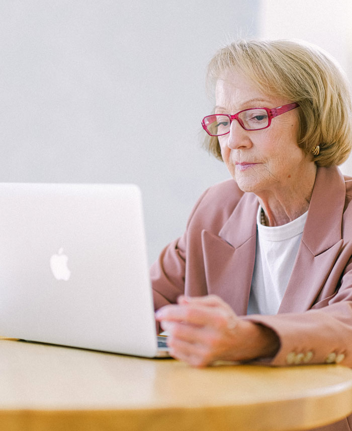 Elderly woman in pink glasses at home, using a laptop to avoid boredom.