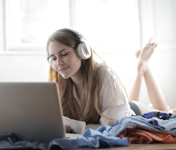 Woman enjoying music with headphones, lying down and using a laptop at home to avoid boredom.