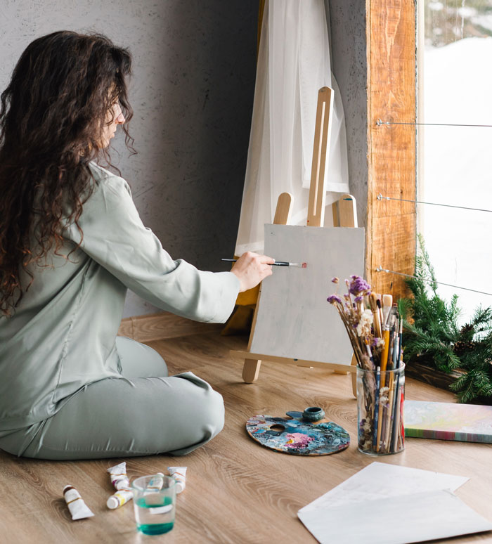 Woman painting on canvas at home, surrounded by art supplies.