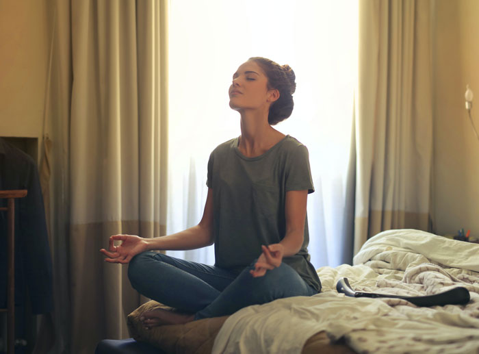 Woman meditating at home, sitting cross-legged on a bed, surrounded by cozy curtains and soft light.