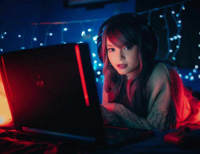 Woman at home gaming on laptop, wearing headphones, with lights in the background for a fun indoor activity.
