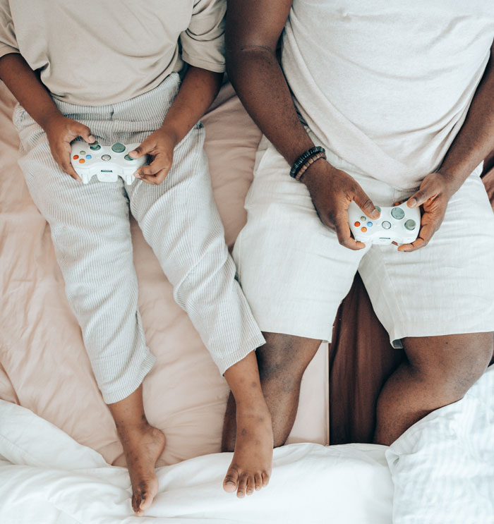 Two people sitting on a bed playing video games at home to avoid boredom, using white controllers.