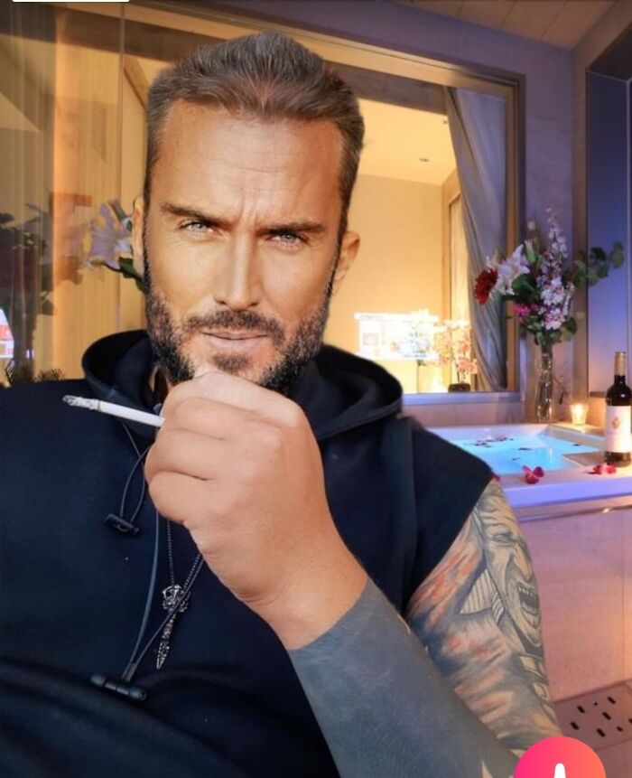 Man with tattooed arm holding cigarette, posing in a well-lit room with flowers and a bathtub, Instagram reality theme.