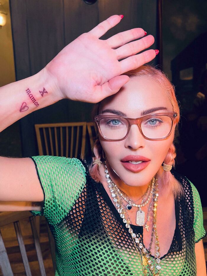 Person wearing glasses and layered necklaces posing with hand raised, illustrating Instagram reality and fake pics online.