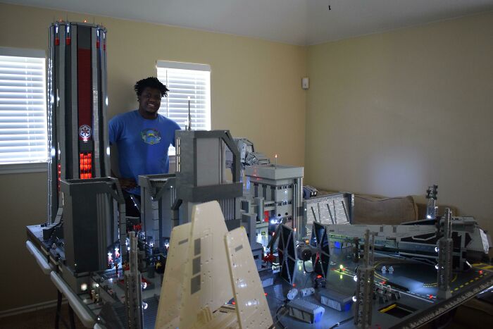 Man in blue shirt showcasing incredible LEGO builds with illuminated detailed structures in a well-lit room setting