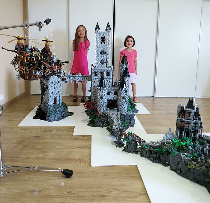 Two girls standing behind an incredible LEGO castle build with detailed stonework and surrounding structures indoors.