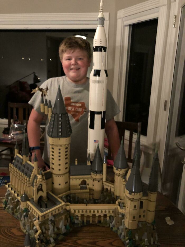 Young boy smiling behind incredible LEGO builds of a castle and rocket displayed on a wooden table indoors.