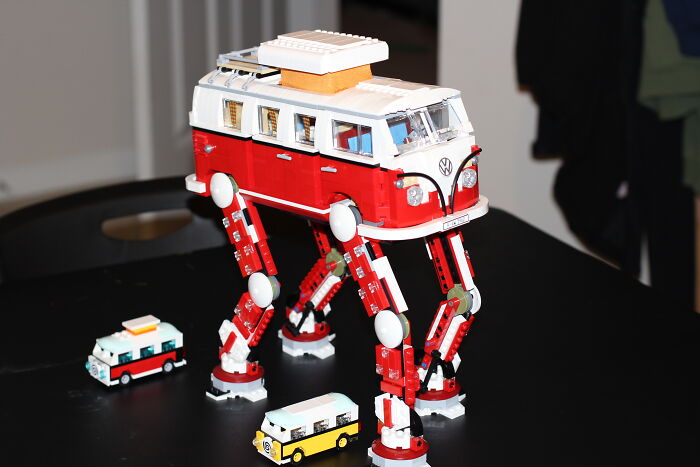 LEGO builds featuring a red and white Volkswagen bus with mechanical legs surrounded by smaller LEGO vehicles on a black table.