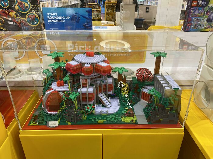 Intricate LEGO builds displayed in a glass case featuring a detailed futuristic house with surrounding trees and plants.