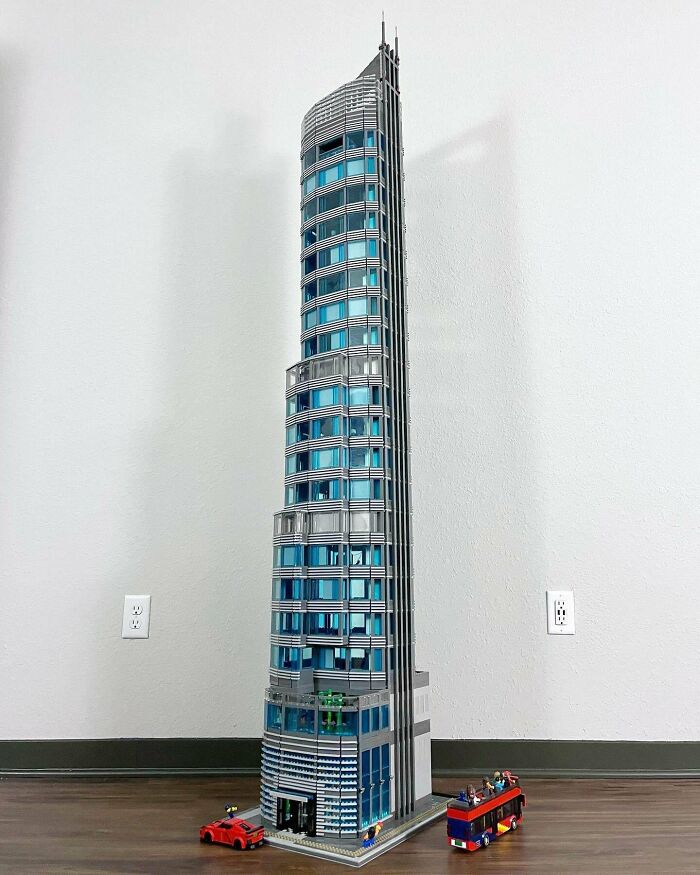 Detailed LEGO building of a modern skyscraper with blue windows and miniature vehicles on a wooden floor.