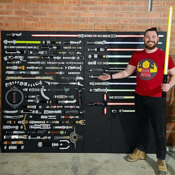 Man holding yellow LEGO lightsaber, standing next to a display of intricate LEGO builds of various weapons on a wall.
