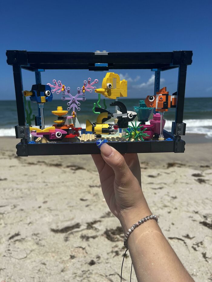 Hand holding colorful LEGO builds of fish and underwater scene with ocean and beach in the background.