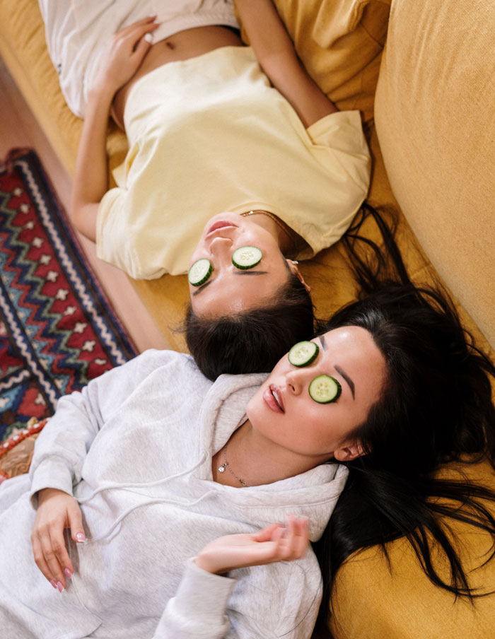 Two women relaxing at home with cucumber slices on eyes, lying on a mustard couch.