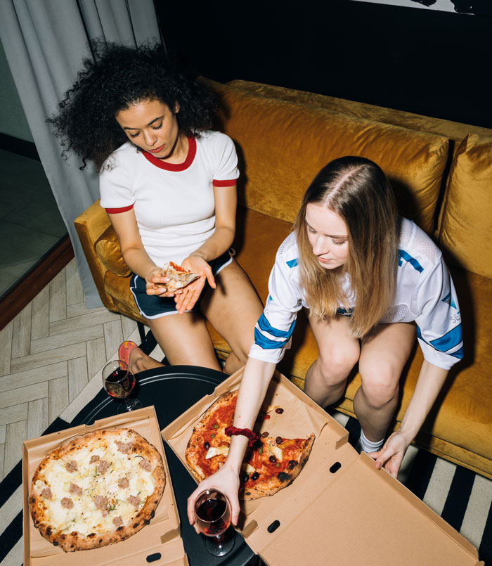 Two women enjoying pizza and wine at home, having fun together on a cozy evening.