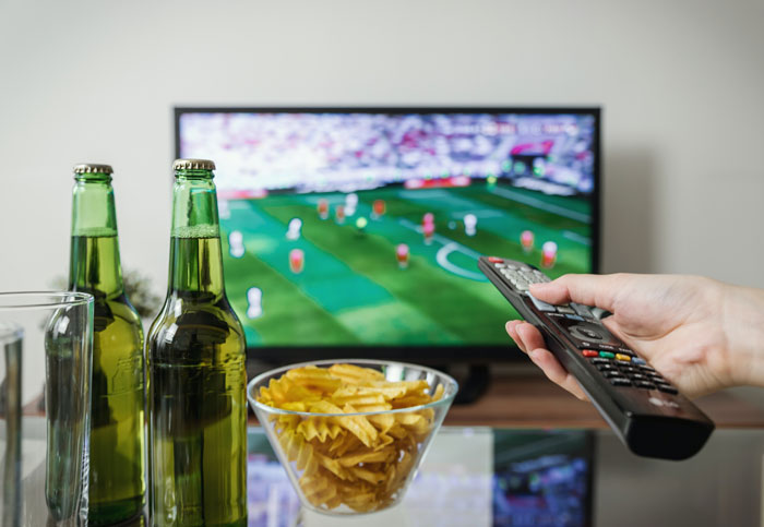 Watching soccer on TV, hand holding remote, chips and beer on table, enjoying fun things to do at home.