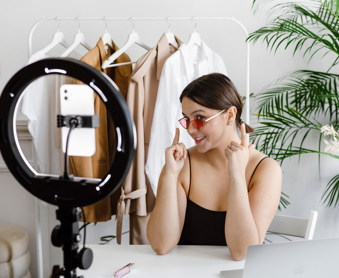 Woman filming a fun video at home with a ring light and smartphone, clothes on rack in background.