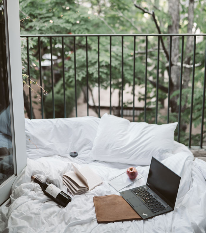Cozy home setup with a laptop, book, and wine on a bed by a balcony, perfect for avoiding boredom.