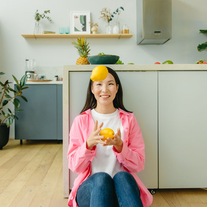 Woman juggling citrus fruits at home, sitting in a kitchen and smiling, enjoying fun indoor activities to avoid boredom.