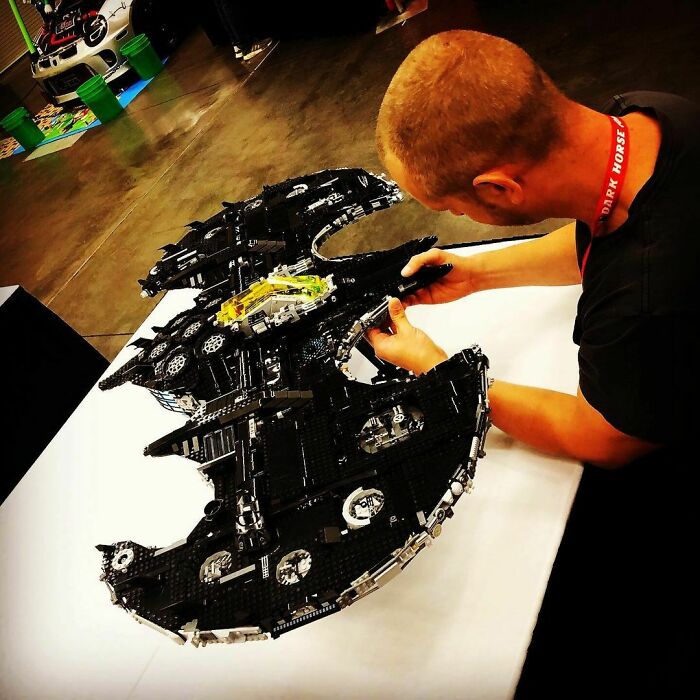 Man assembling an incredible LEGO build resembling a detailed black spacecraft at an indoor event with other LEGO displays nearby