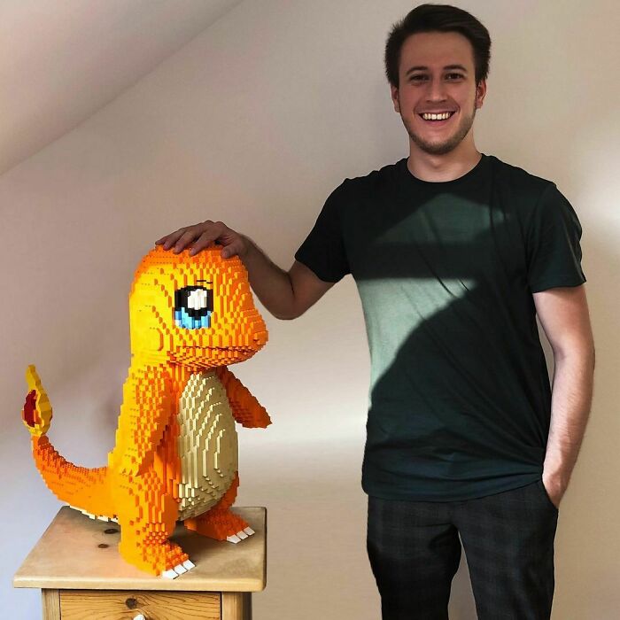 Young man smiling next to an incredible LEGO build of a large orange character with a flame tail on a wooden stool.