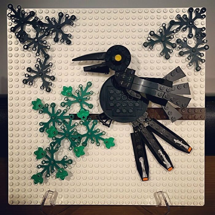 Intricate LEGO build of a black bird perched on a branch with green leaves and decorative elements on a white baseplate.