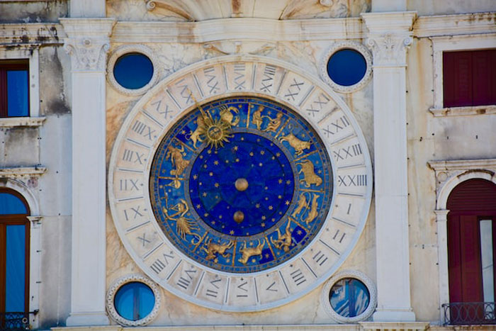 Ornate zodiac clock on historic building facade, representing time and astrology with gold accents and Roman numerals.
