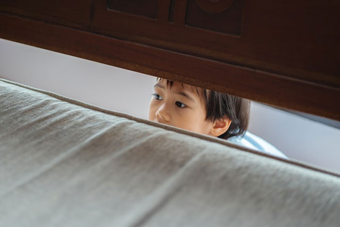 Child peeking nervously from behind a bed, evoking creepy stories about children's imaginary friends and chilling experiences.