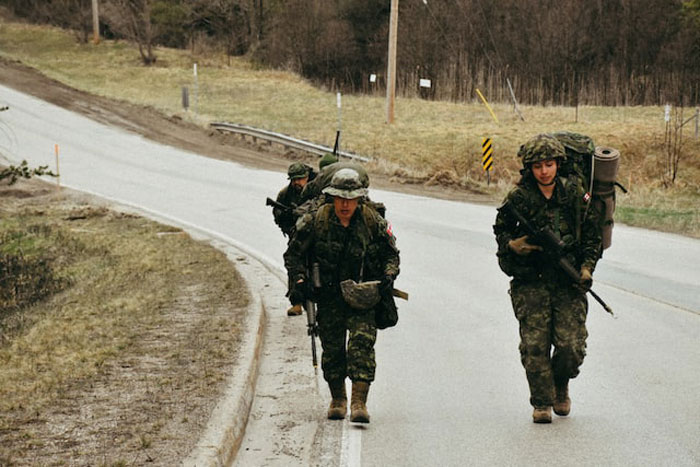 Three soldiers in full combat gear walking along a rural road surrounded by grass and trees in overcast weather.