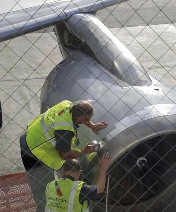 Two workers in safety vests tape an airplane engine, ignoring OSHA safety regulations, at an airport.