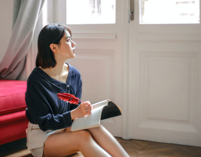 Young woman holding red feather and notebook, sitting on floor by window, reflecting on curious facts in art classes.