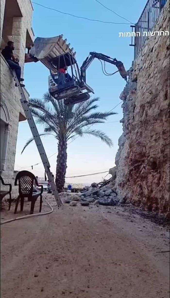 A person in a loader bucket hangs high above the ground, defying OSHA safety regulations, with a ladder beside.