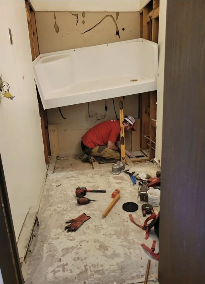 Worker under a precariously placed bathtub, ignoring OSHA safety, in a makeshift construction setup with tools scattered.