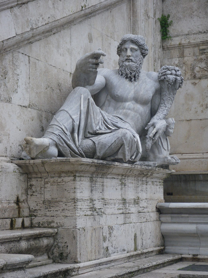 Marble statue of a reclining bearded man holding a cornucopia on a stone pedestal in an outdoor art setting.