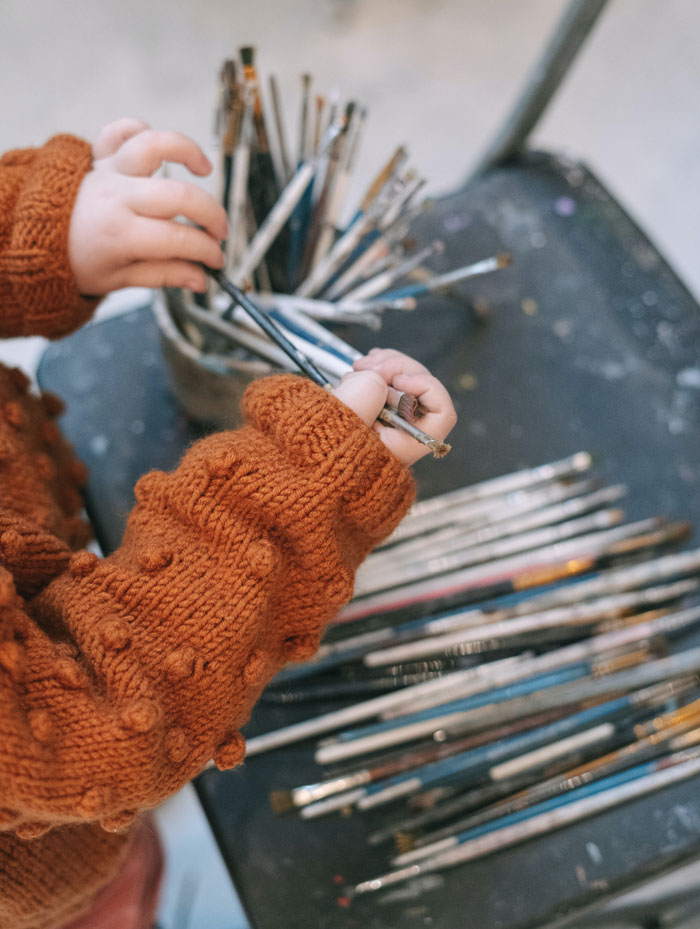 Child in a brown sweater choosing paintbrushes from a container, highlighting curious facts in art classes.