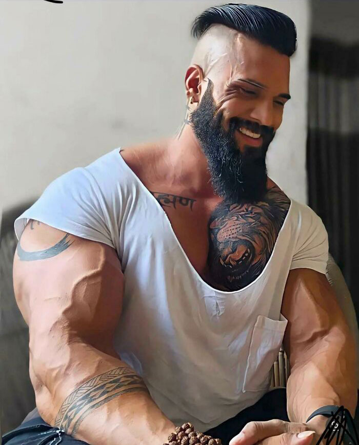 Muscular man with tattoos and a beard wearing a loose white shirt, posing with exaggerated arm size in an Instagram reality photo.