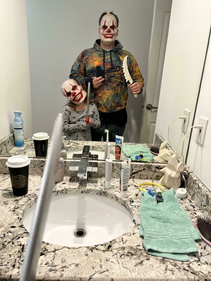 Parent and child in Halloween masks taking mirror selfie holding toy weapons, capturing kids making parents laugh.
